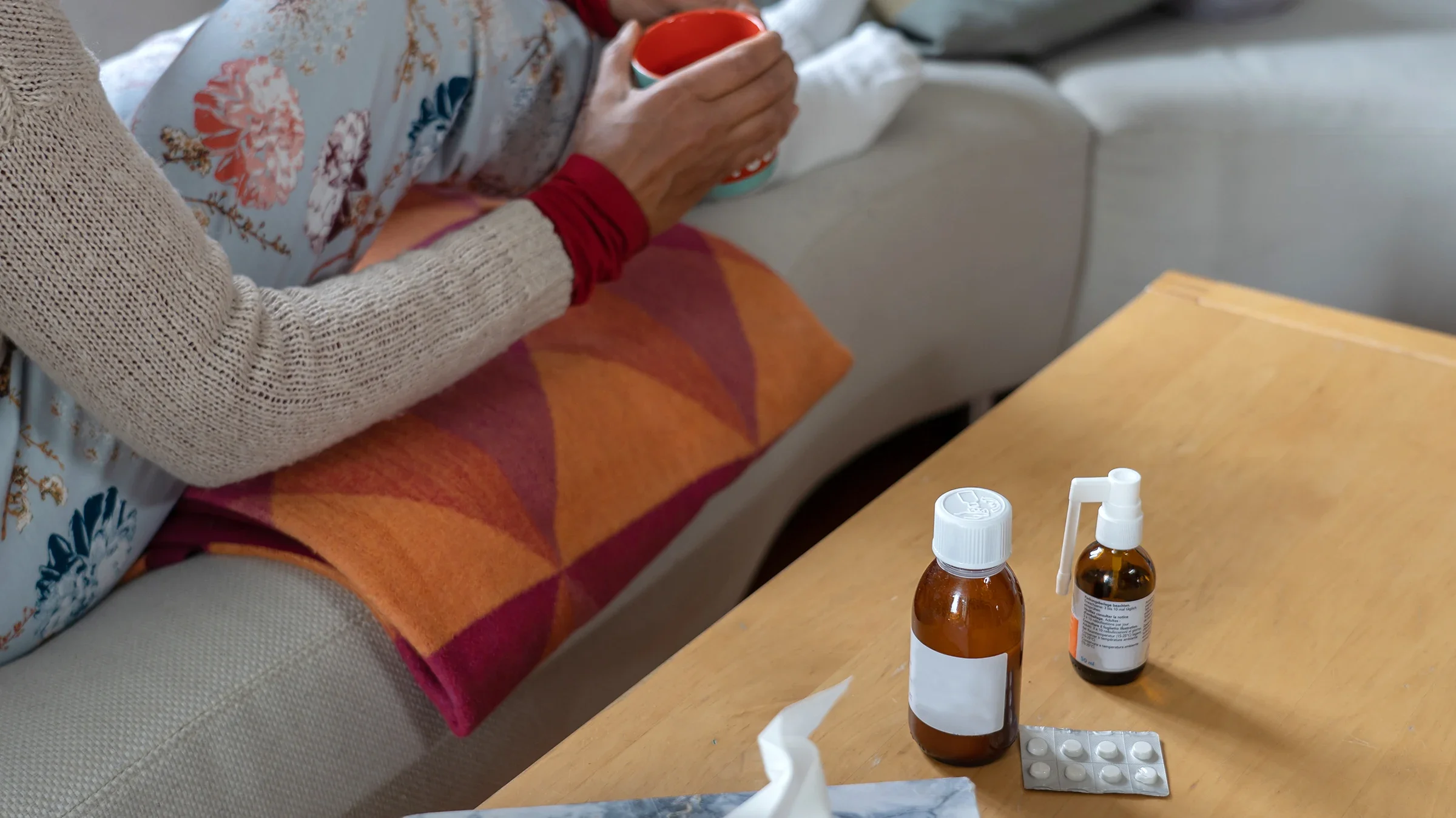 Close-up of a woman sitting on the couch holding a cup of tea with cold medication sitting on the coffee table next to her, only her legs and hands are in view.
Claudine Silaho Weber-Hilty/iStock via Getty Images