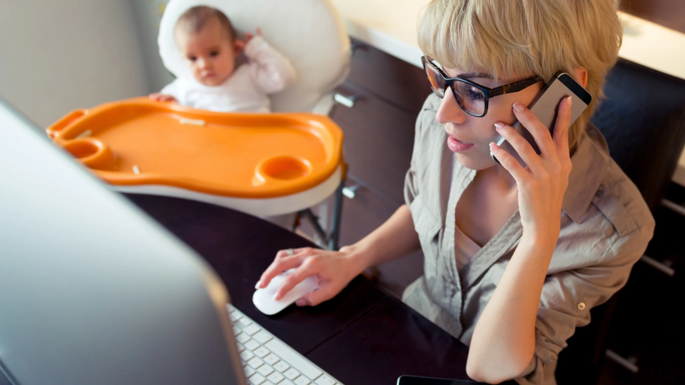 A parent on a phone call while using their computer, with their baby next to them.