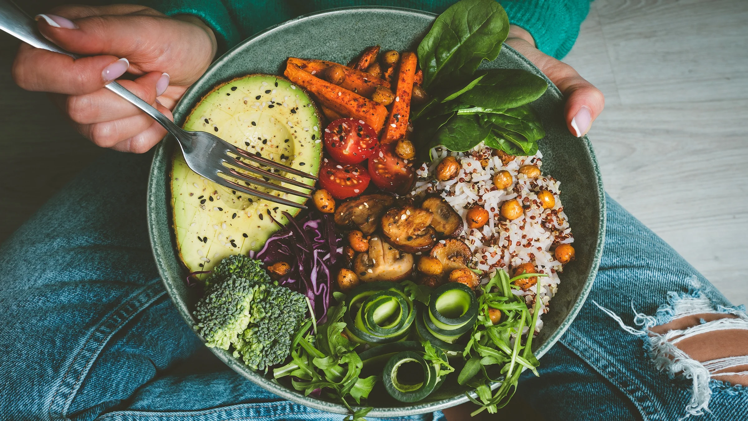 A woman eats bowl of fresh vegetables, grains, and mushroom.
