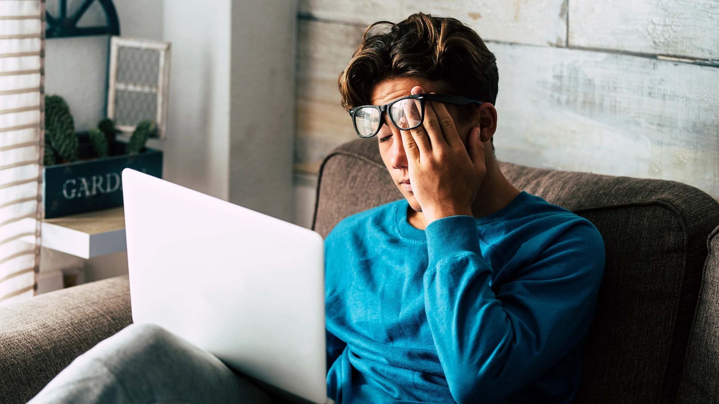 Young man looking tired while working on his laptop. He is pushing his glasses up to his forehead while rubbing his eyes.