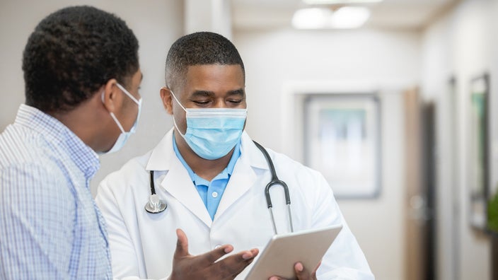 Doctor explaining test results to a patient. The doctor is pointing to the tablet that they are reading from. Both are wearing face masks.