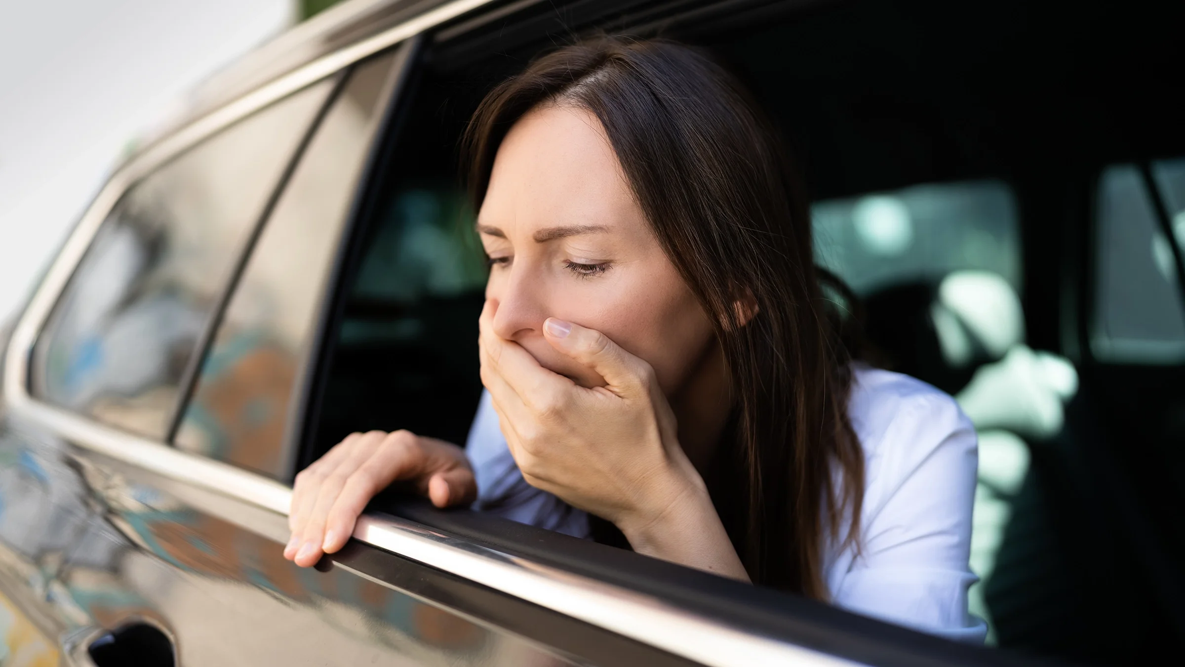 A woman experiencing nausea leans her head out of a car window.