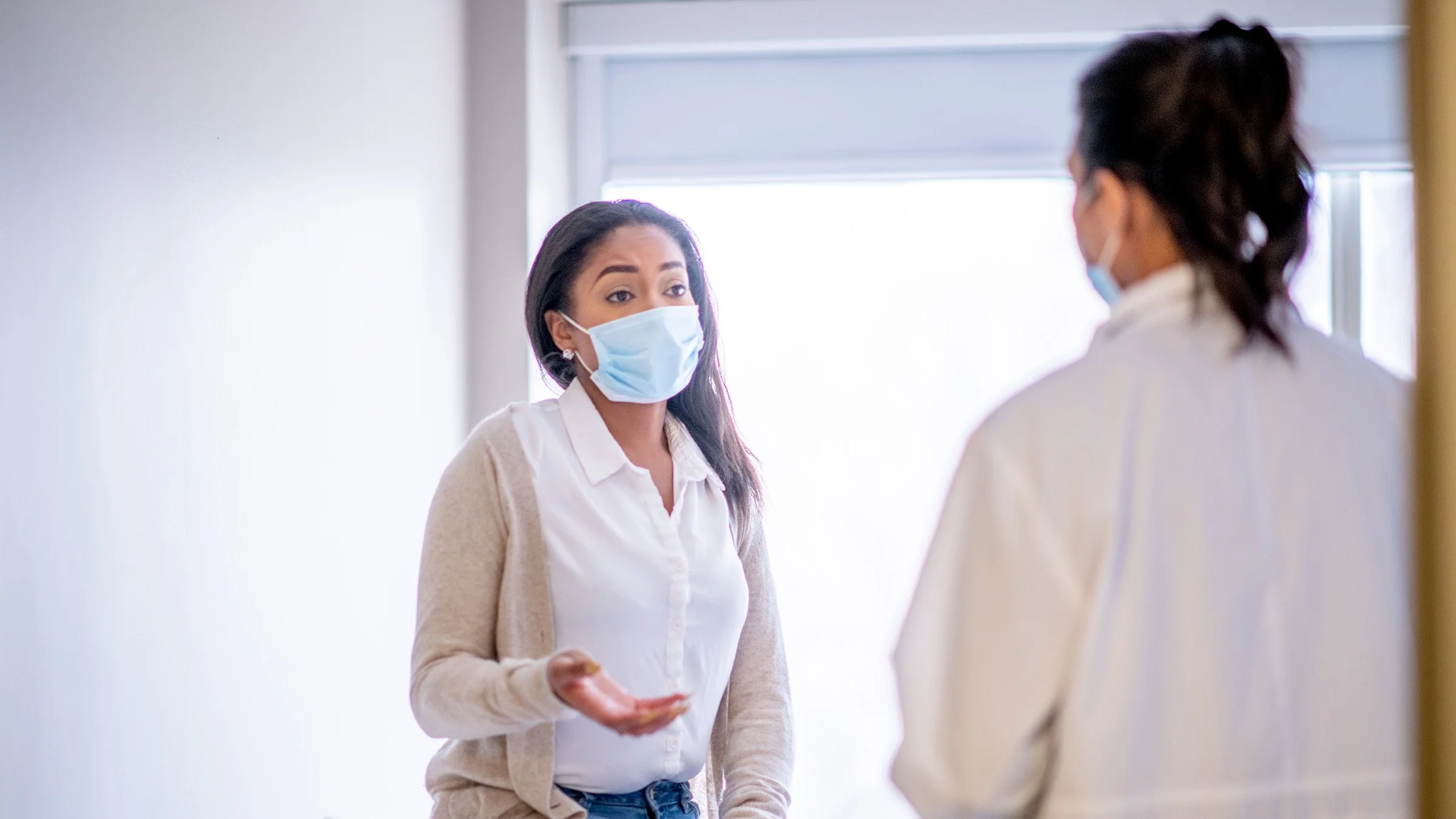 Young woman in the exam room at her doctor's office. She is wearing a medical face mask and has her hand out like she is asking a question to the doctor.