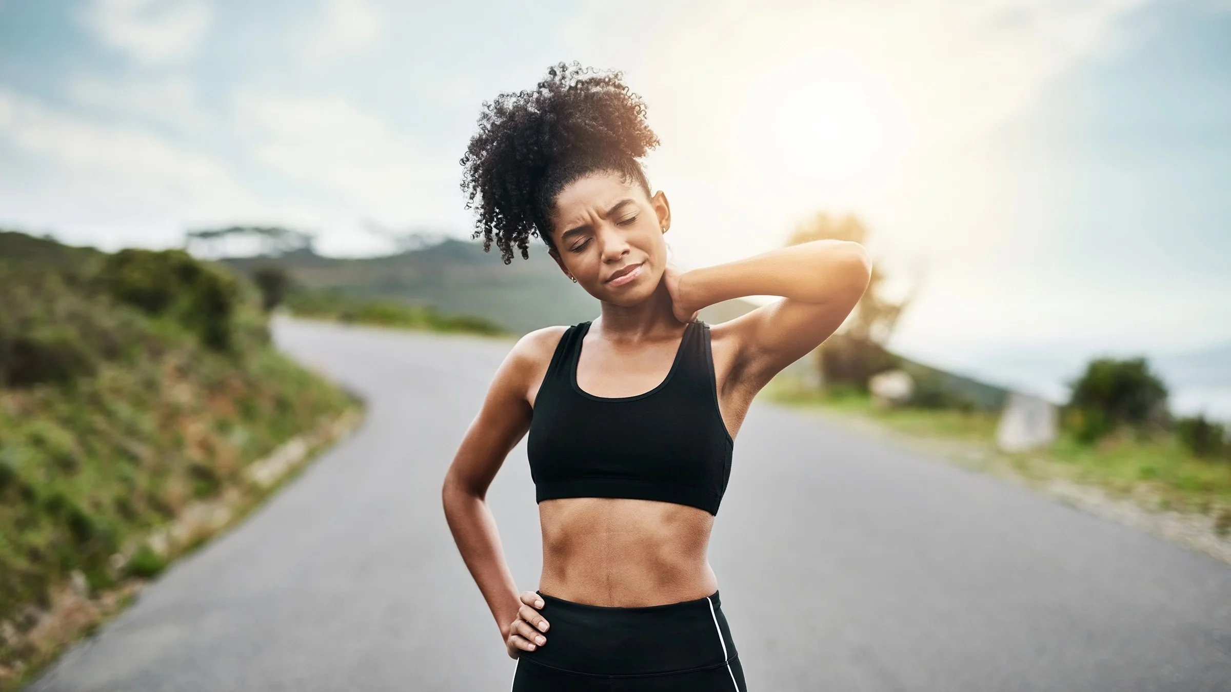 Portrait of a woman stopping on her run. She is rubbing her back and neck like it is sore.