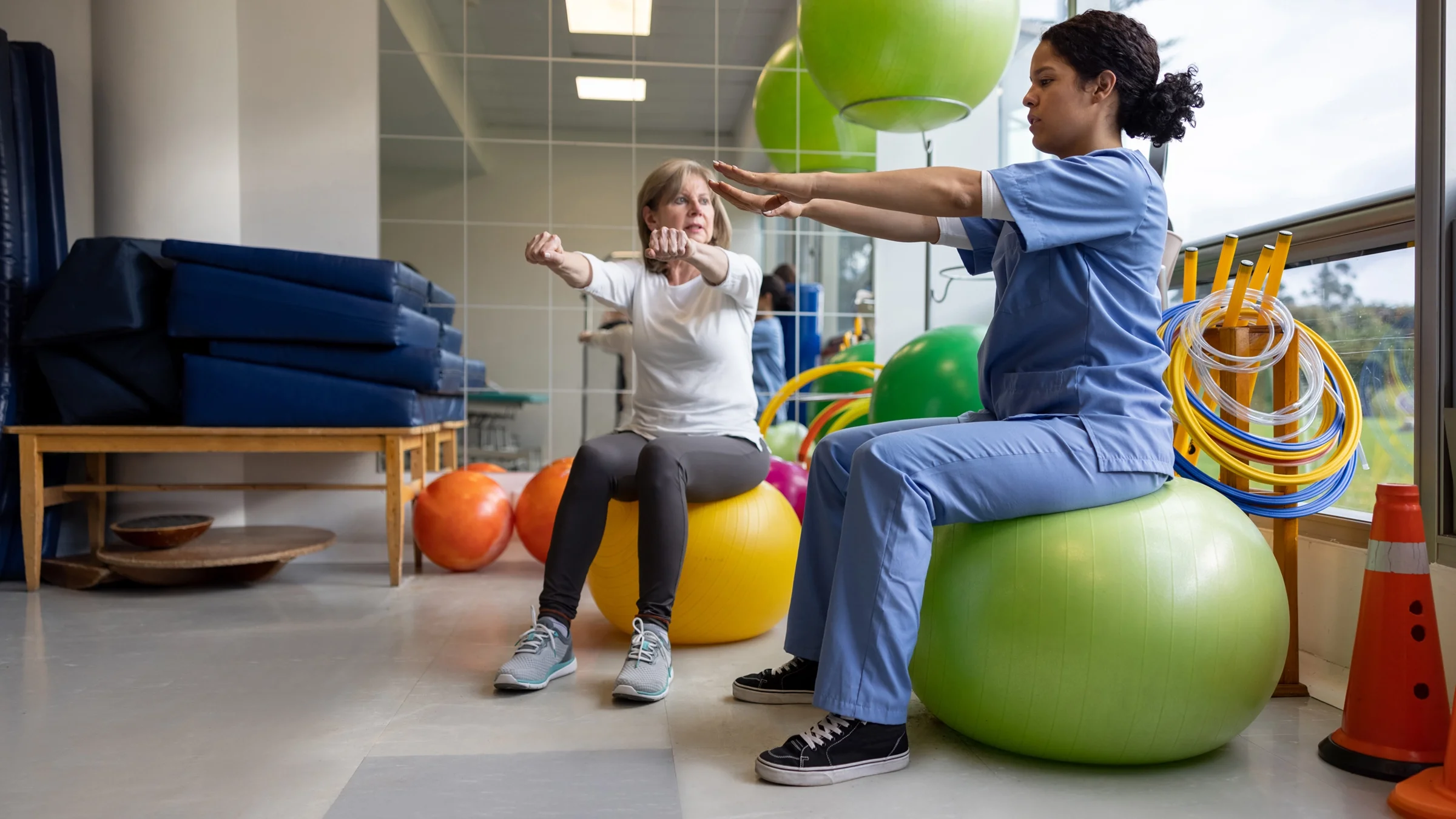 Senior woman at occupational therapy with nurse. They are sitting on exercise balls doing arm stretches.