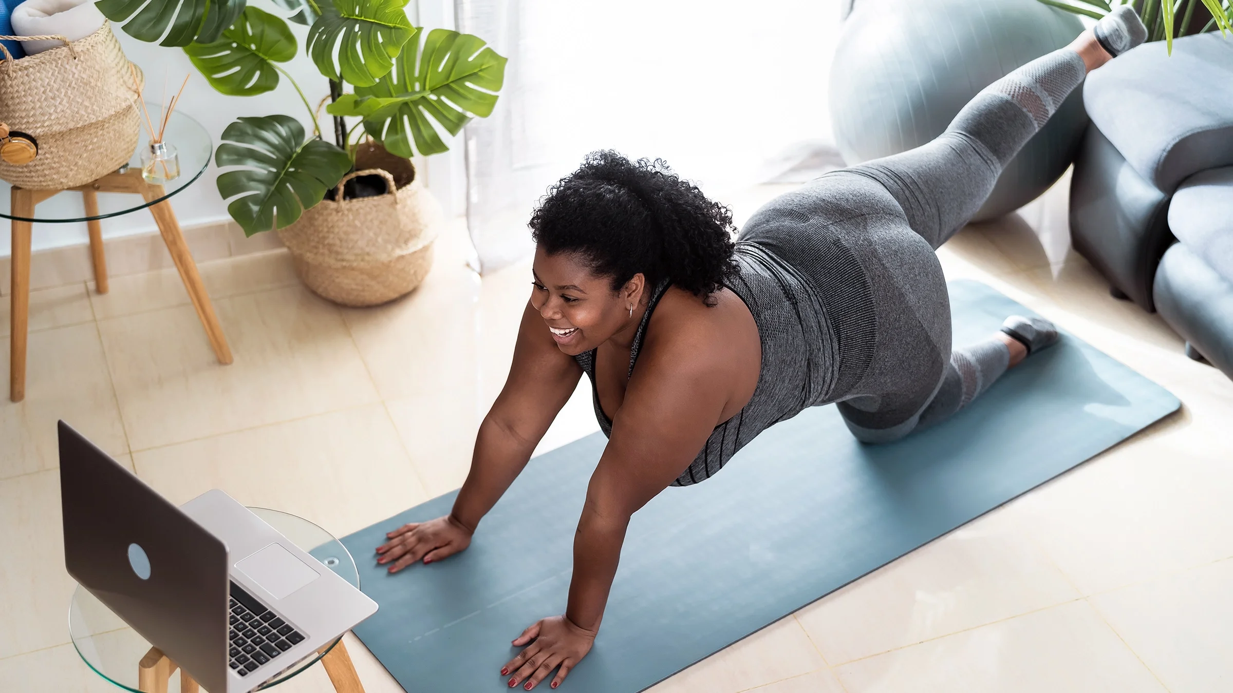A woman is following along with a virtual Pilates class on her laptop at home.