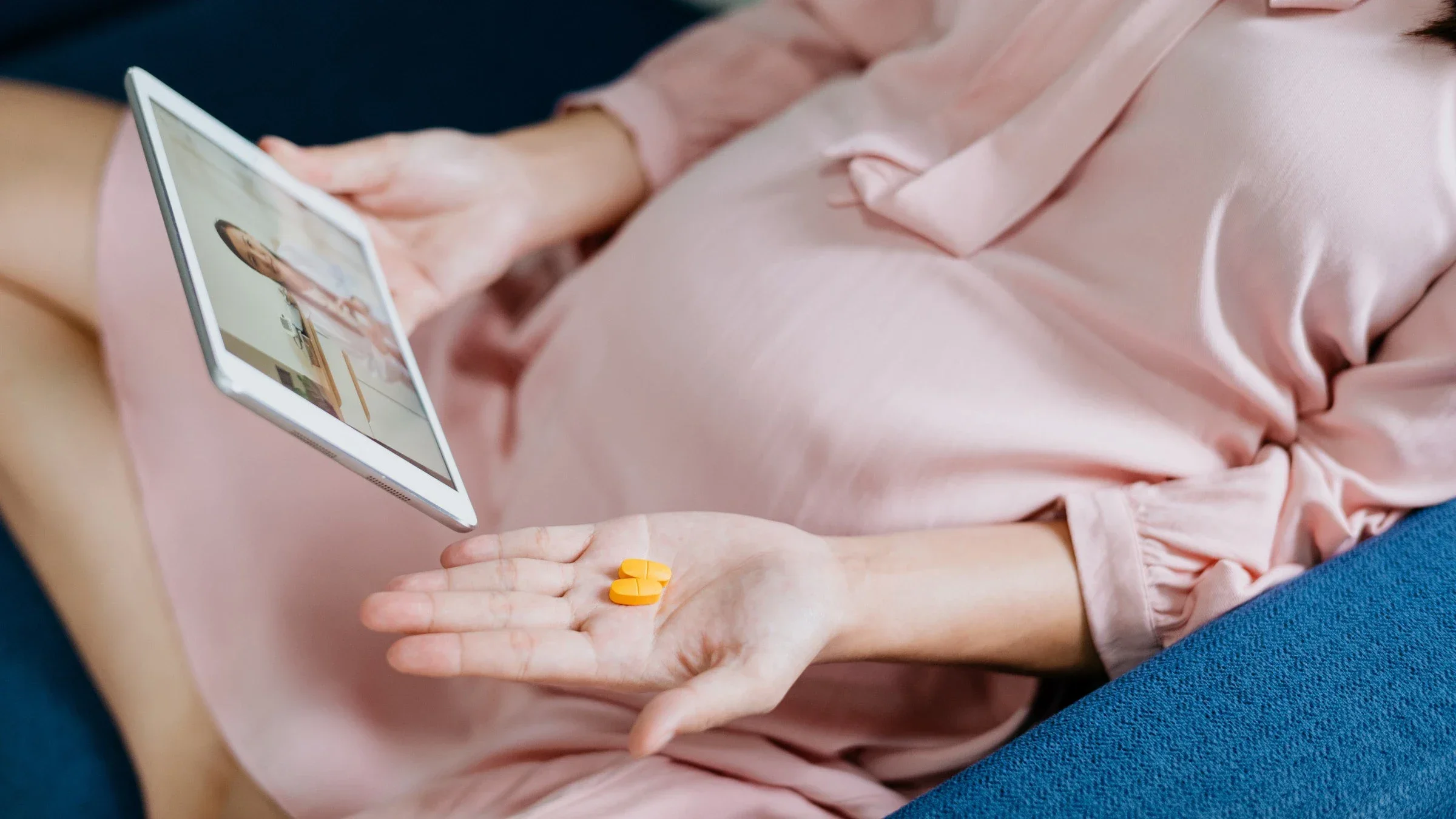 Pregnant woman sitting cross legged on the couch on a telehealth appointment with her doctor. She has two orange pills in one hand and the tablet in the other.
hxyume/E+ via Getty Images