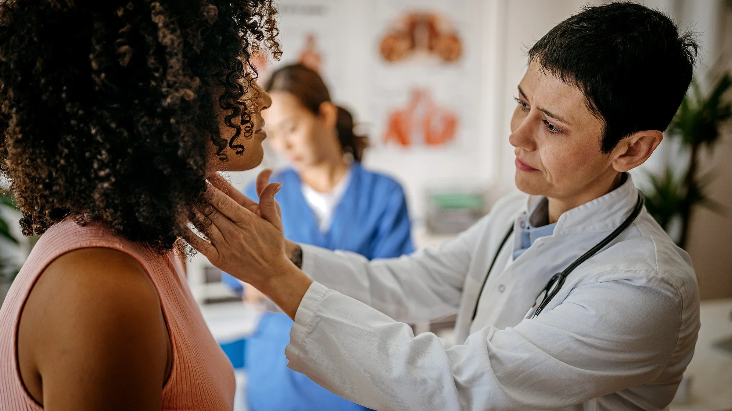 Patient with curly hair getting her thyroid examined by a doctor. The nurse is in the background out of focus as well.
mixetto/E+ via Getty Images