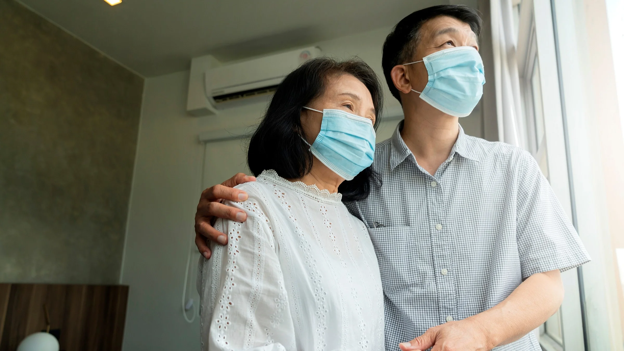 Portrait of an older Asian couple holding each other as the look out their window. They are wearing blue medical face masks.
