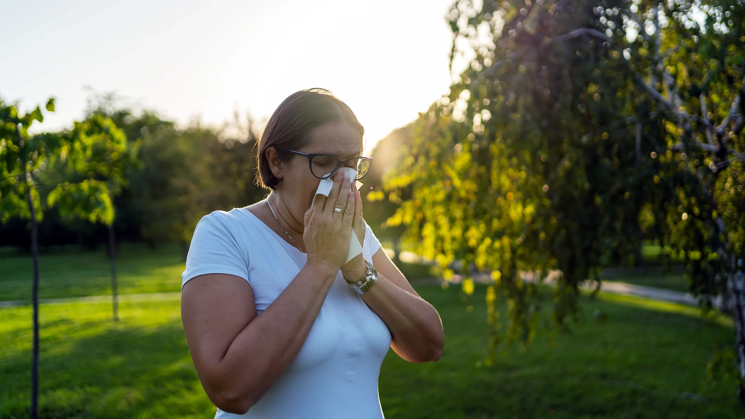 A woman with allergy symptoms blows her nose outside.