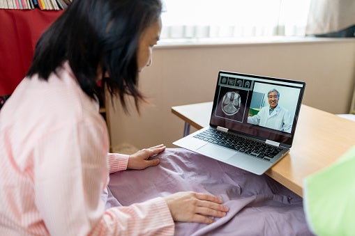 A woman sitting in bed with her laptop on a telehealth visit with her doctor looking over her MRI scan on screen.