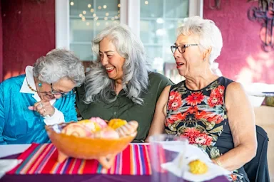 Three elderly women sitting at an outdoor table smiling and laughing together.
adamkaz/iStock via Getty Images