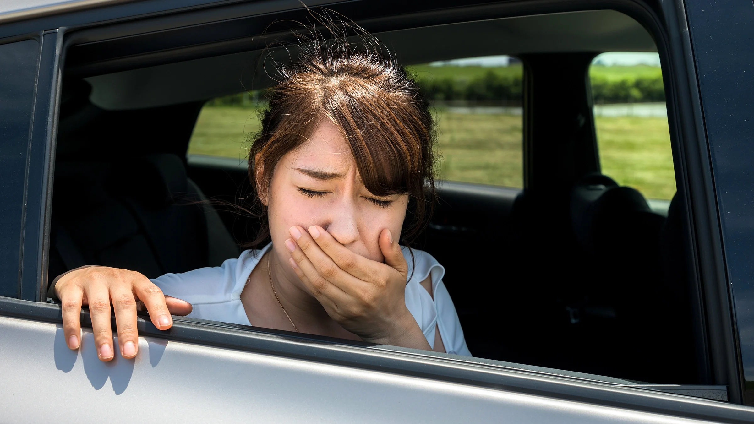 A woman is leaning out of the car window. She is covering her mouth and looks like she is experiencing motion sickness.