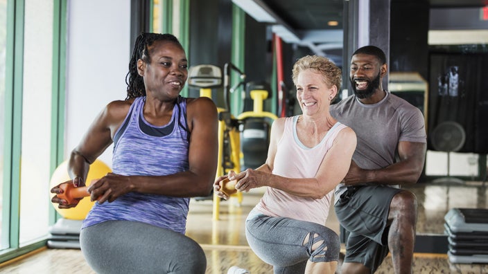 A group of people are shown at the gym doing lunges while holding weights.