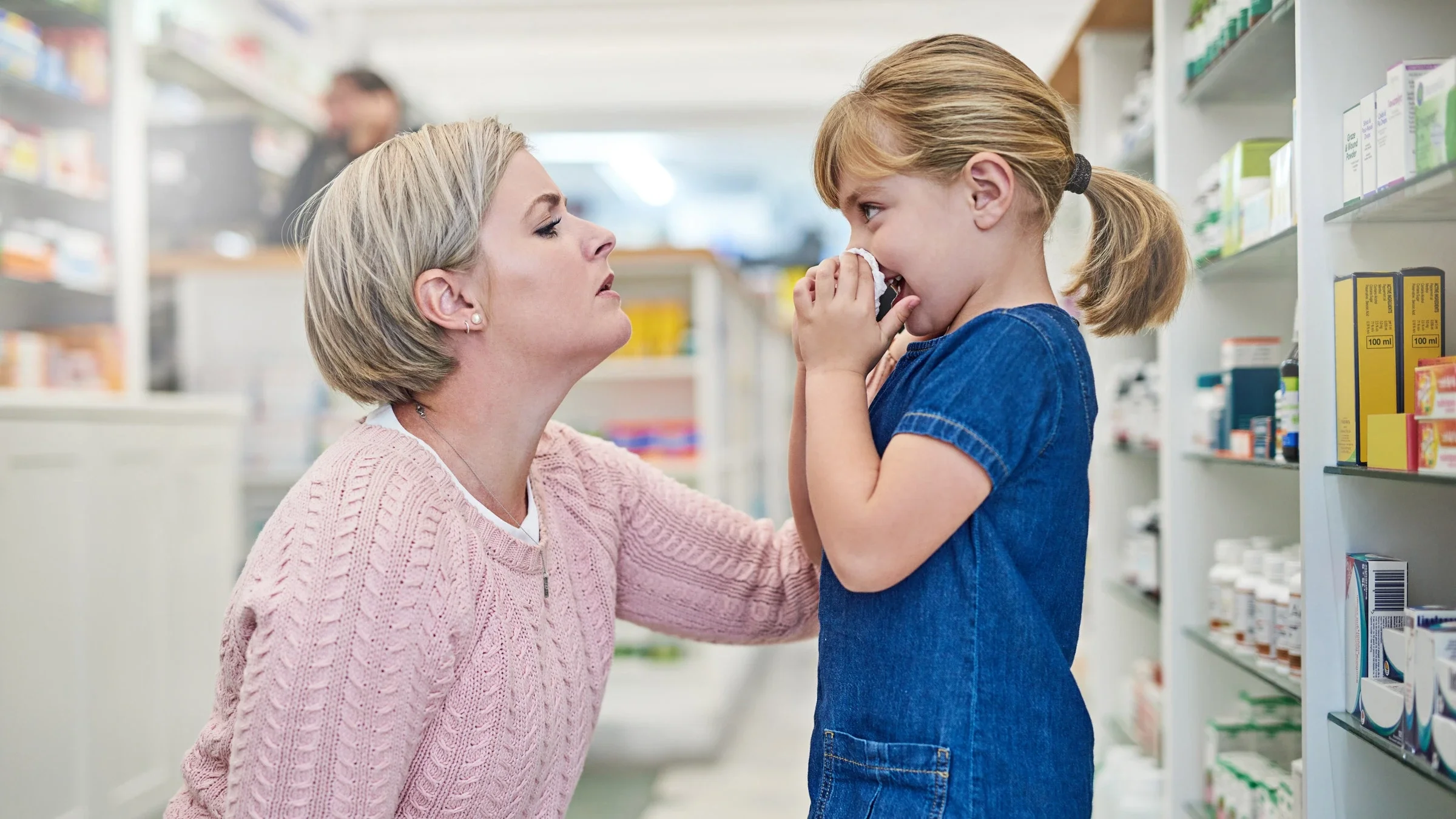 A parent comforting their child who is blowing their nose in a pharmacy.