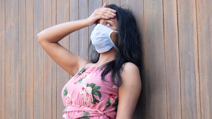 Young woman wearing a face mask and using her hand to check her temperature on her forehead.
