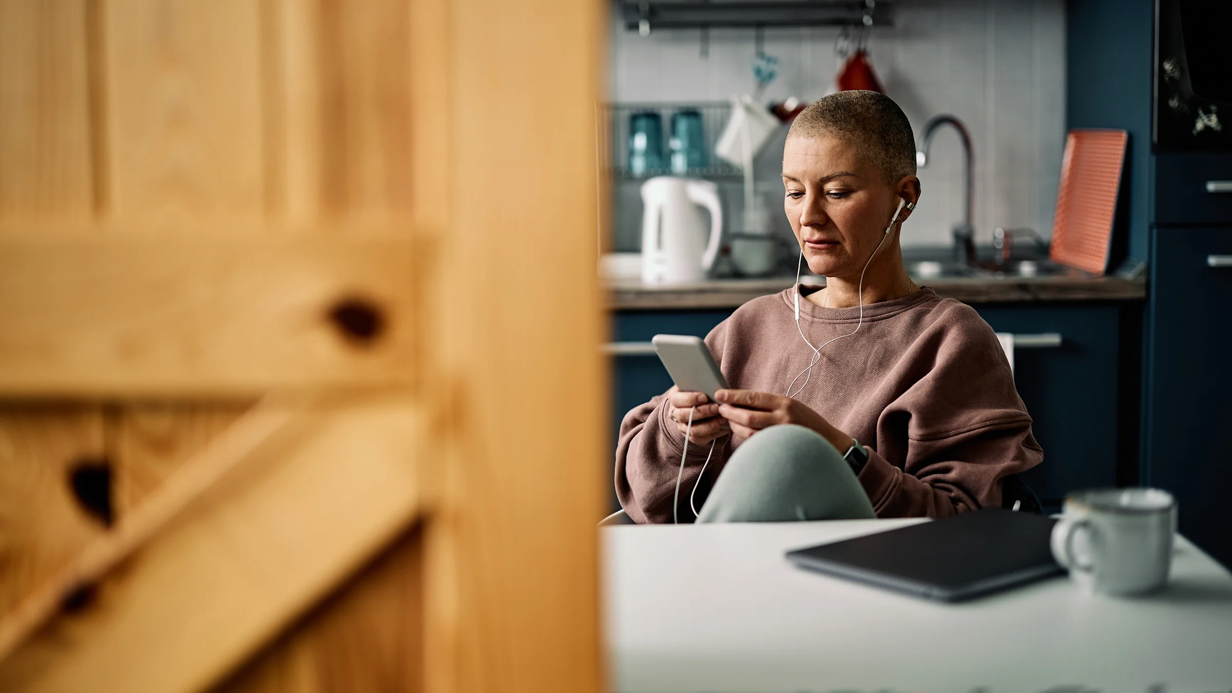 View through a doorframe into a kitchen where we see a bald senior woman is on her phone.