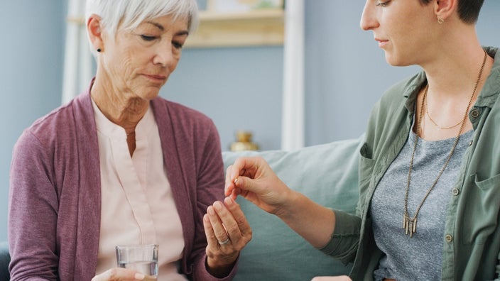 A young woman giving her older mother her medicine. It is a small white pill in between the daughters fingers. The mother is reaching for it and has a glass of water in her other hand.