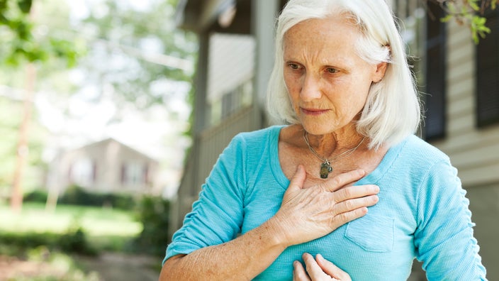 Portrait of an older adult outside, holding a hand to her chest as if she is experiencing chest pain.