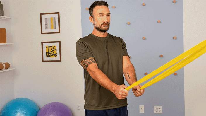 A man demonstrates a shoulder extension exercise by pulling on a resistance band with both arms.