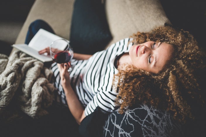 Young woman with curly hair leaning her head back on the couch while drinking wine and holding a book.