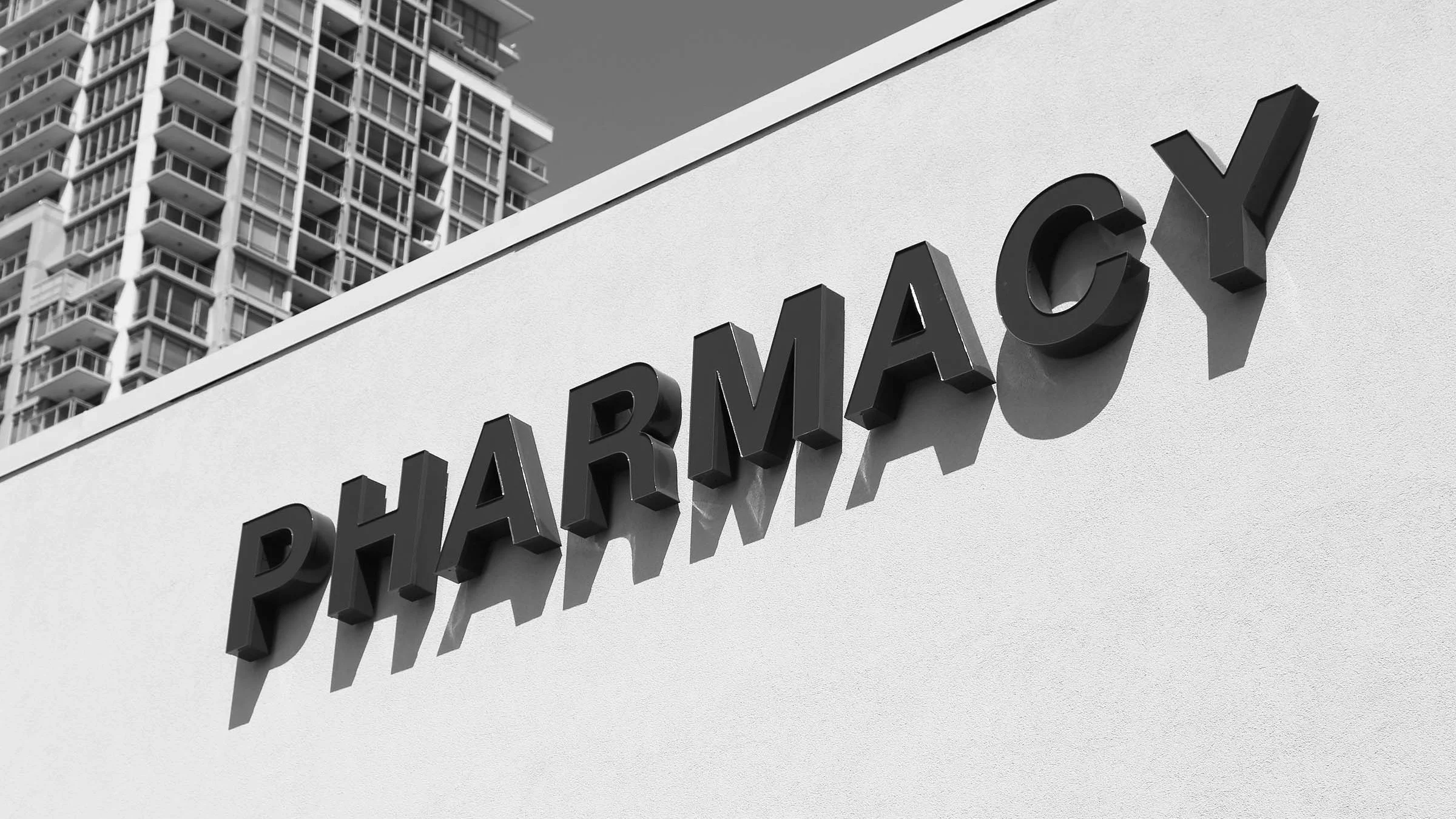 Black and white photo of pharmacy sign on the side of a building with a high rise building in the background.