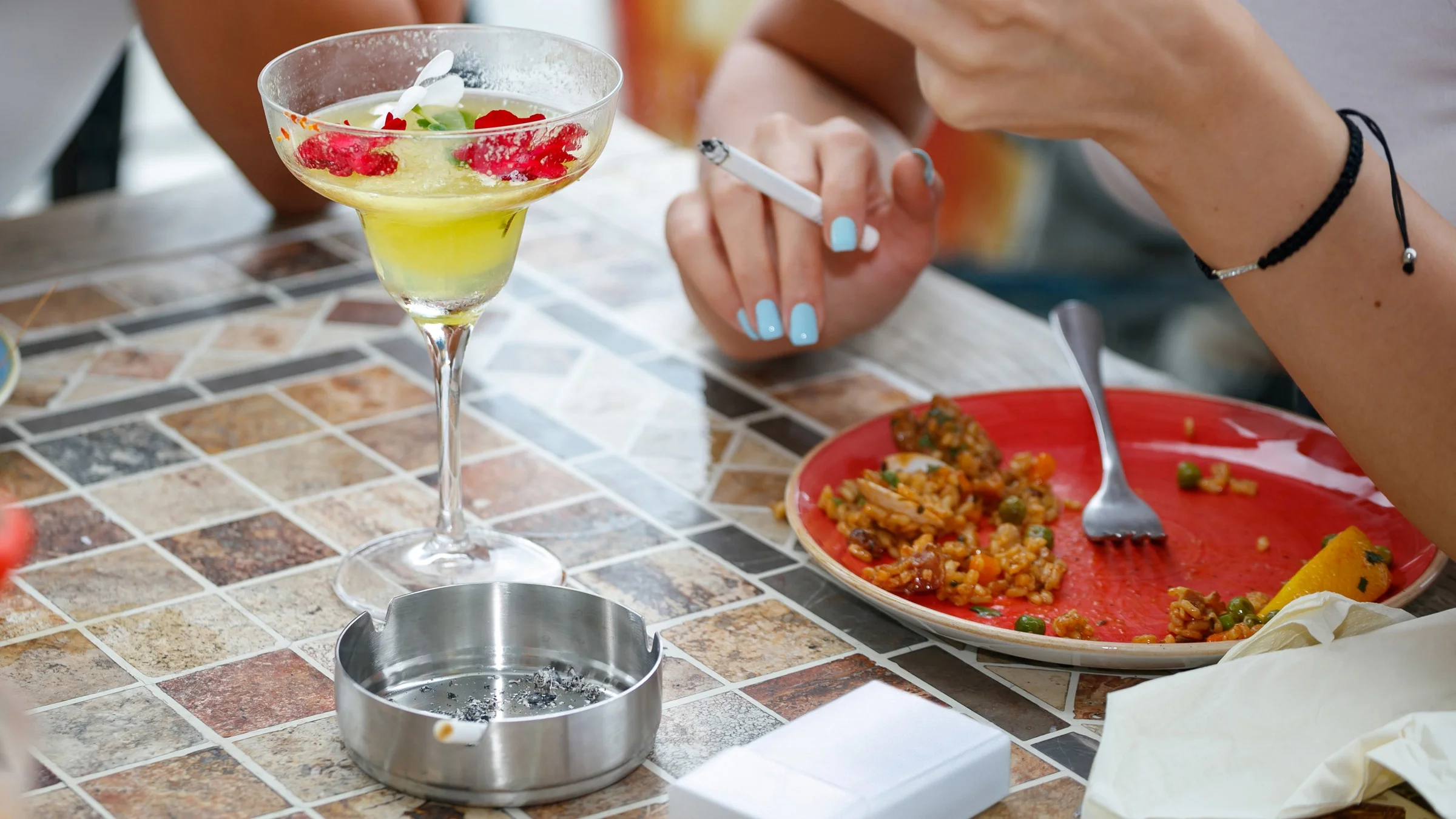 Cropped shot of someone smoking and drinking a cocktail at an outdoor dining establishment.