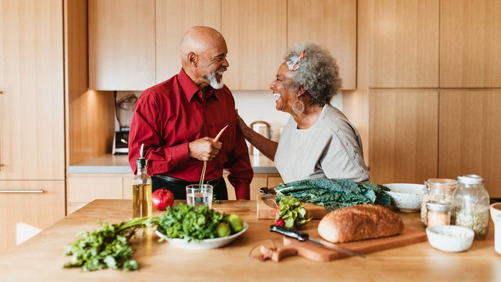 A senior couple is prepping dinner in the kitchen. They are looking at each other and smiling and laughing.