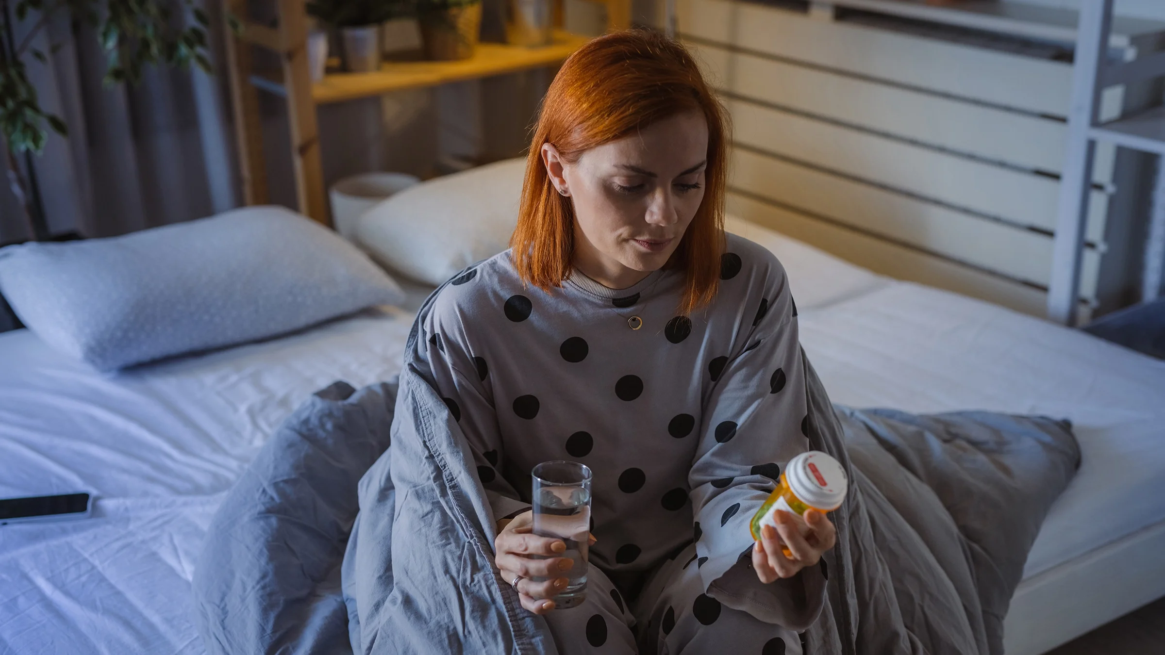 A woman sits on her bed at night holding a medication bottle and a glass of water.