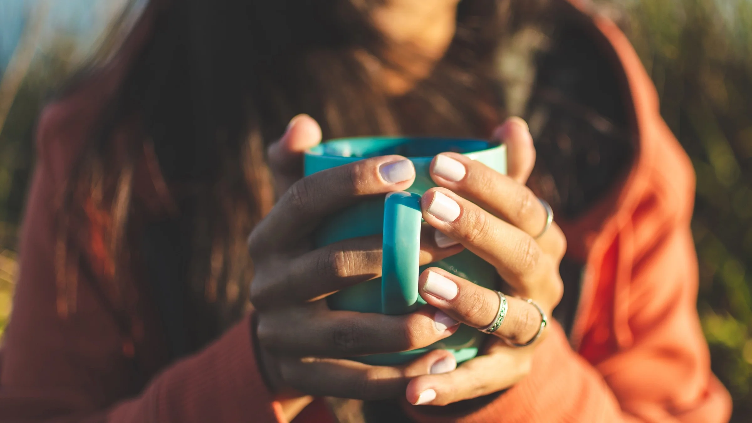Close-up of a person holding a cup of coffee. Both hands are on the teal-colored cup with the handle facing out.