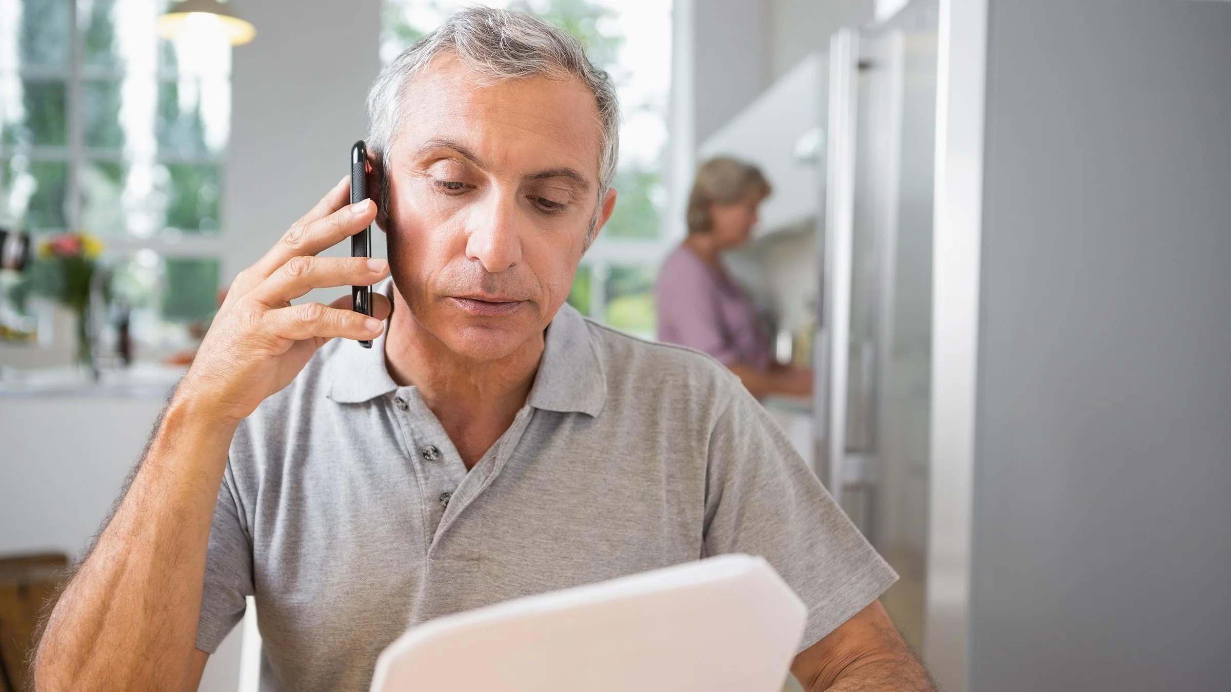 Older man with gray hair on his cell phone talking while he looks at paper work in his hand.