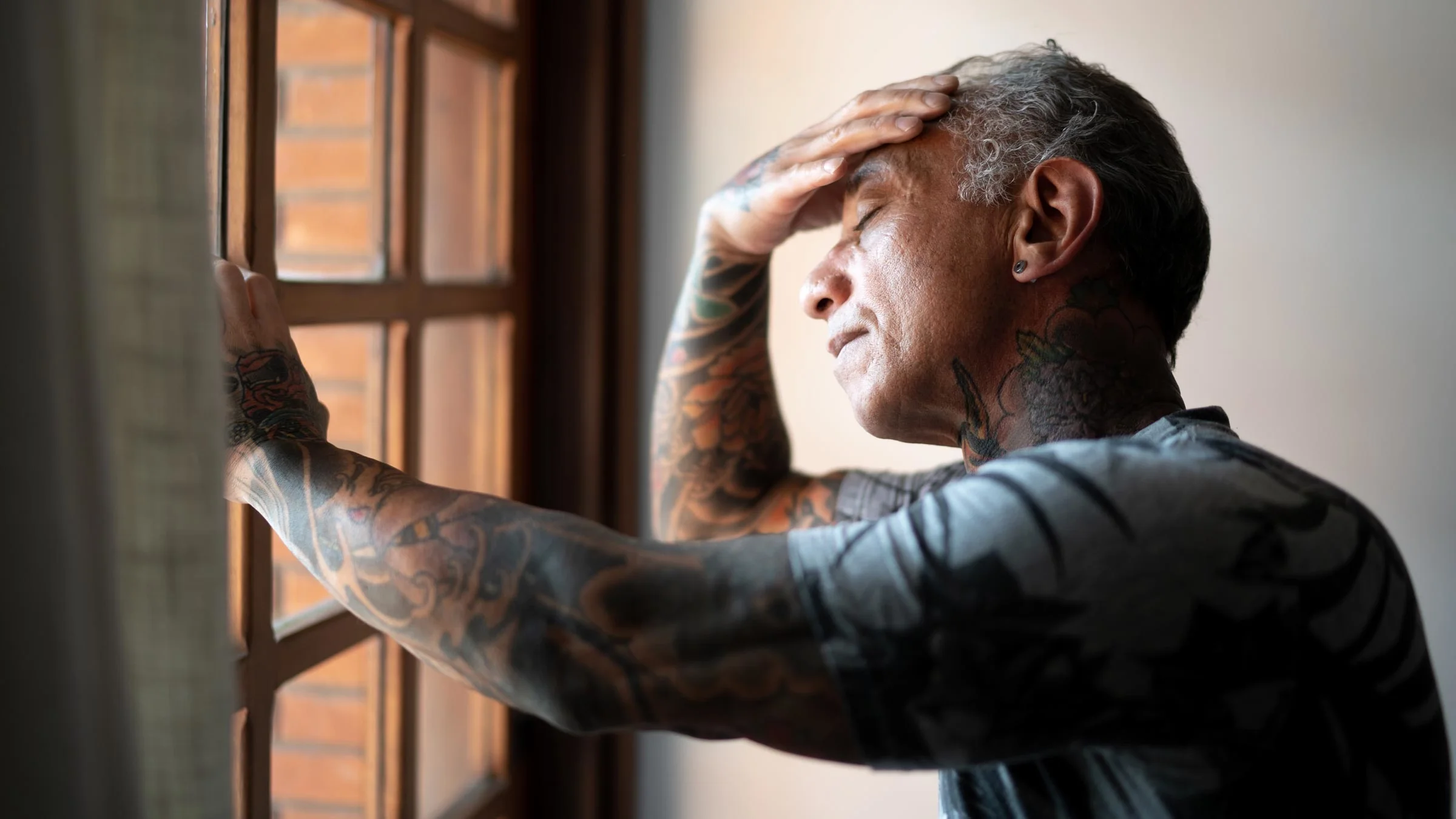 A man stands before a window, clasping a hand to his forehead.