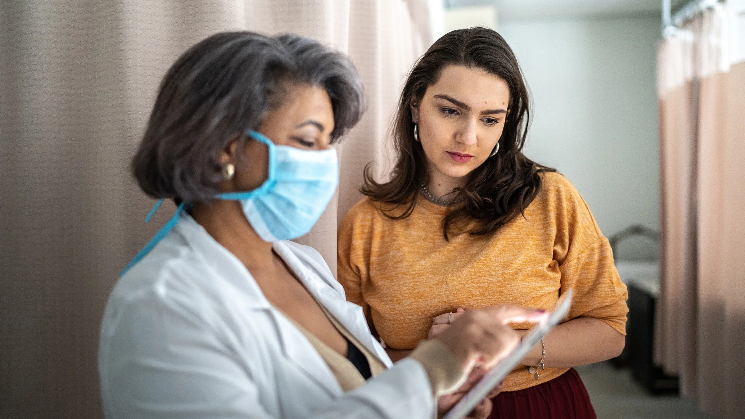 A young woman is having a consultation with her doctor. She is looking over her shoulder at a clipboard.