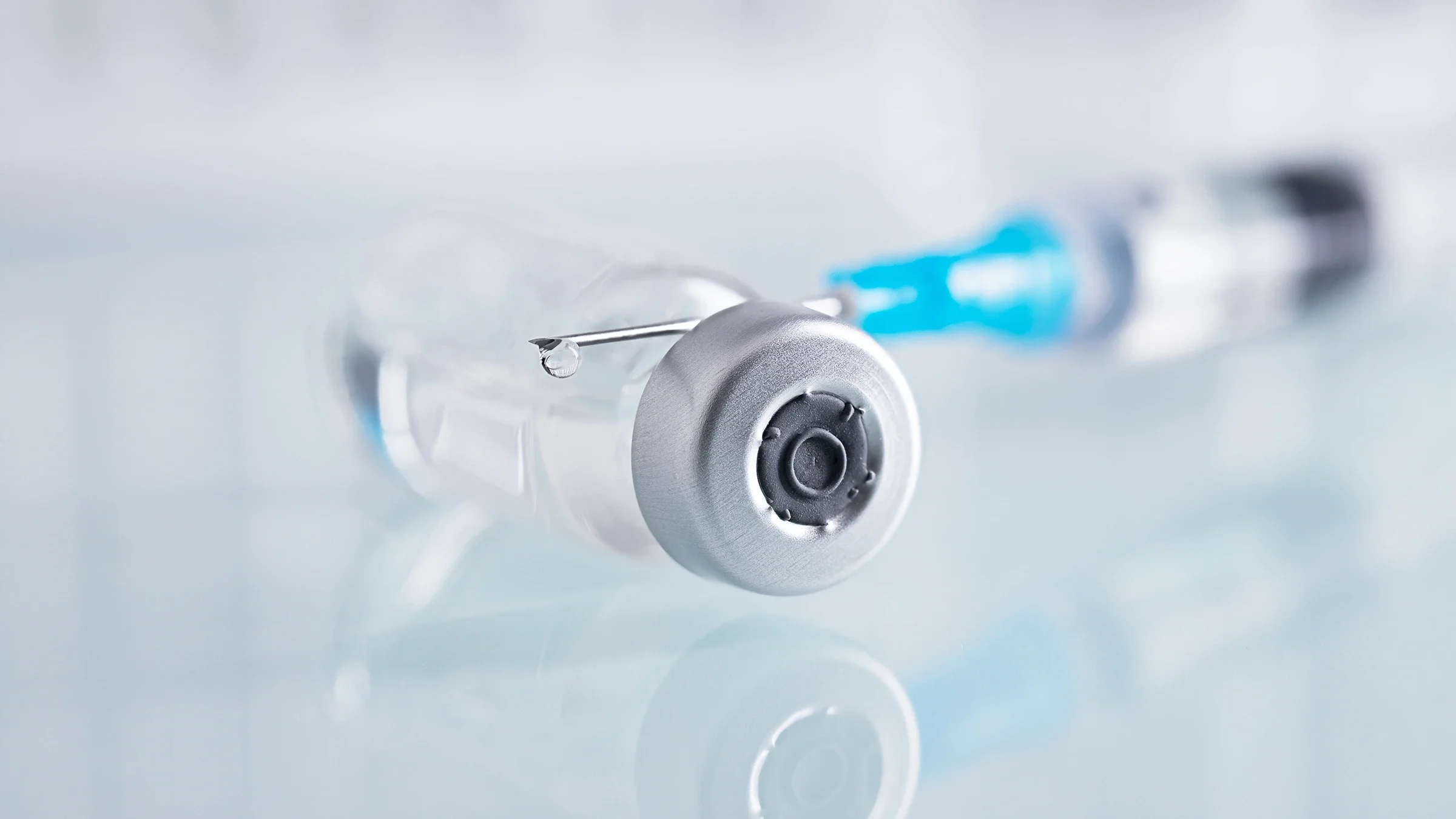 A close-up image shows a syringe and a closed glass vial with clear liquid on a white surface. 