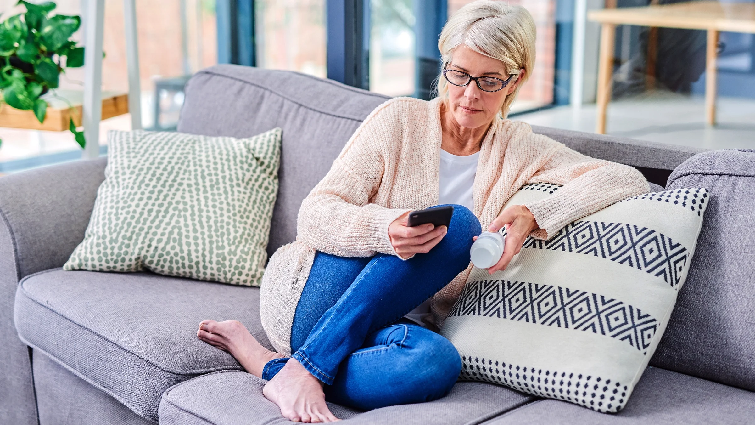 A senior woman sits on a couch using a smartphone and reading the label on a medicine bottle.