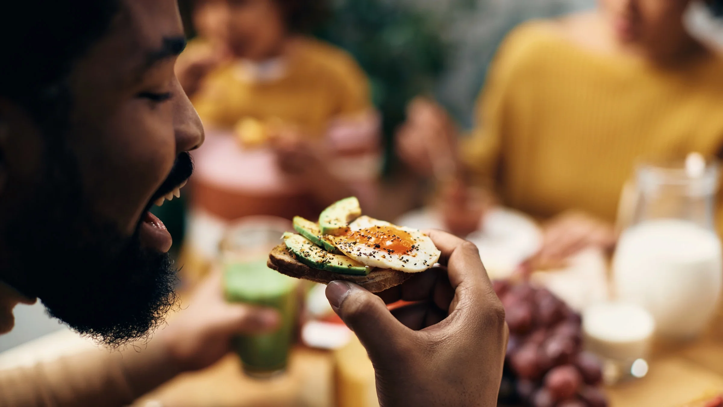 Close-up of a young African-American man about to take a bite of an avocado egg toast, sprinkled with pepper. There are two people wearing yellow shirts in the blurred background.