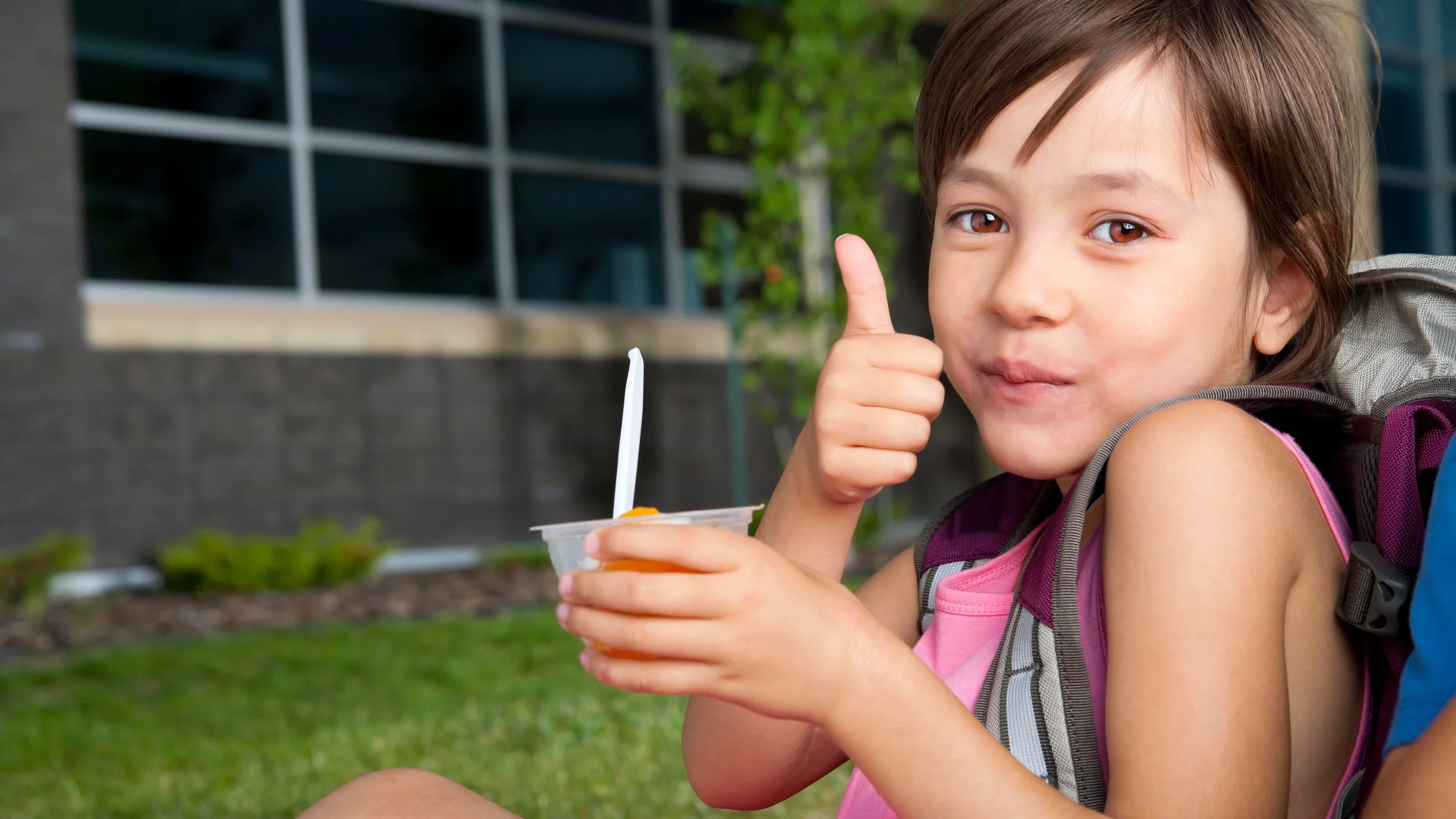 A young student eating a fruit cup at school and giving a thumbs up.