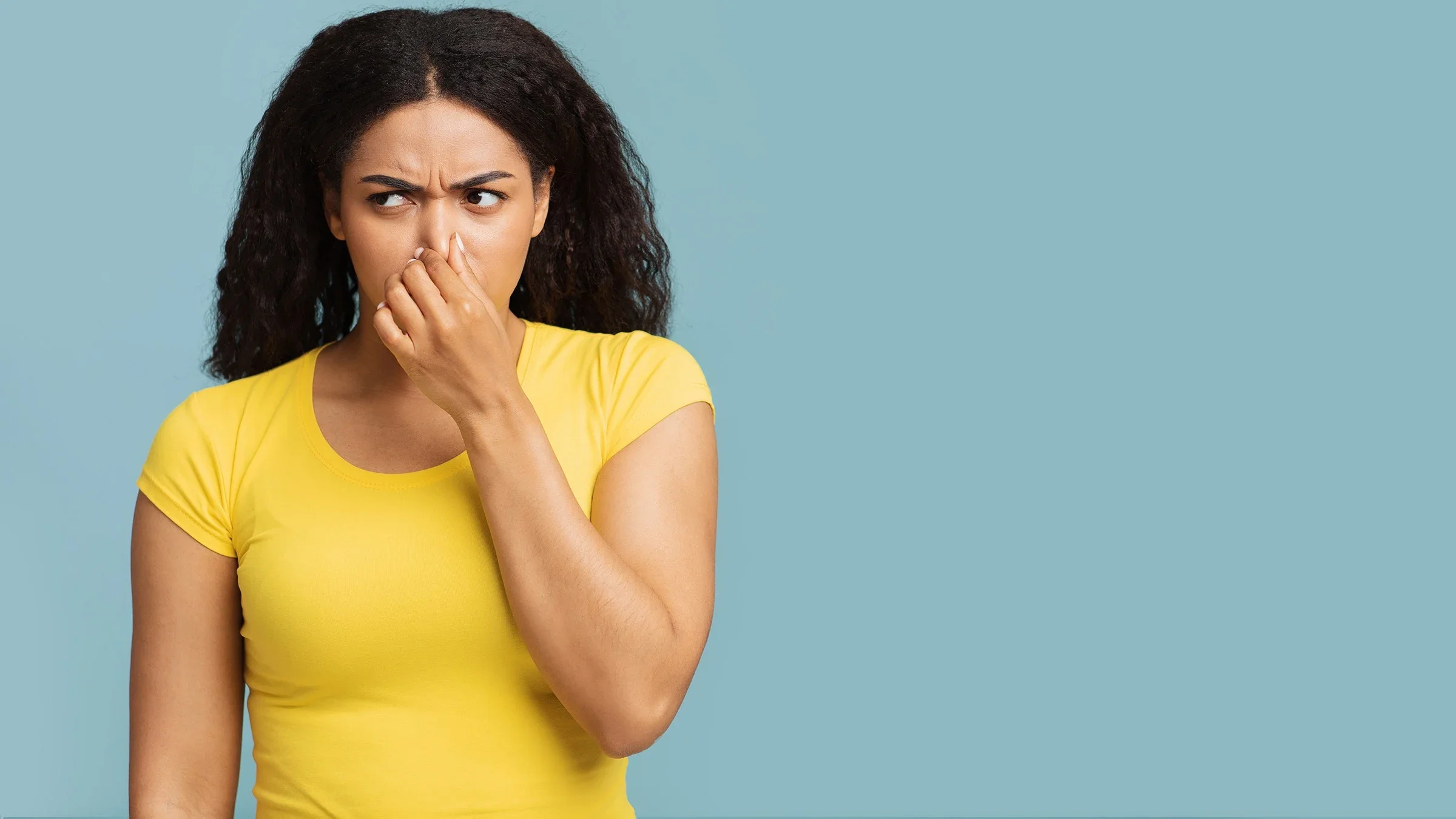Studio portrait of a young woman holding her nose like something smells bad. She is wearing a yellow t-shirt on a light blue background.
Prostock-Studio/iStock via Getty Images
