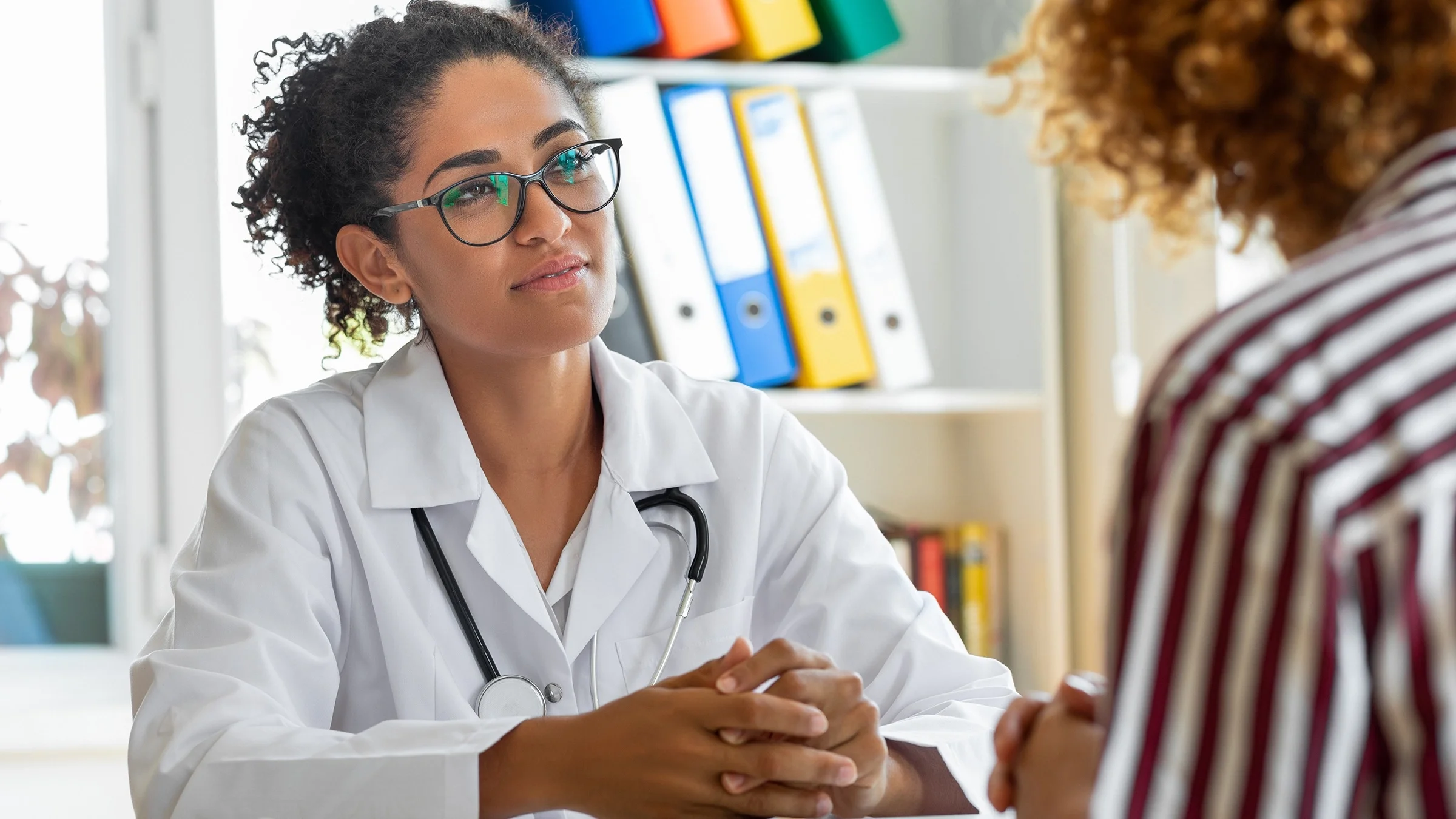 Doctor talking with a patient at her desk. She is looking at the patient with concern and interest. The patient has their back to us and is nondescript.