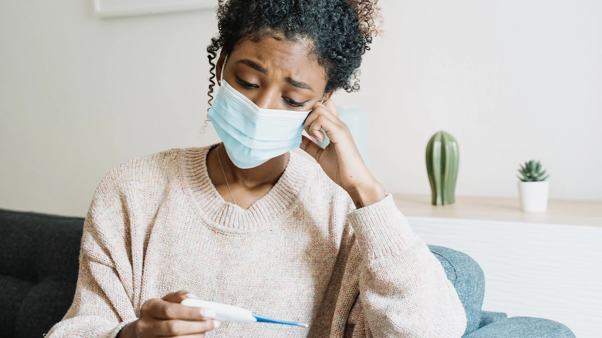 Portrait of young woman sitting on a couch looking at thermometer saddly with blue medical face mask on