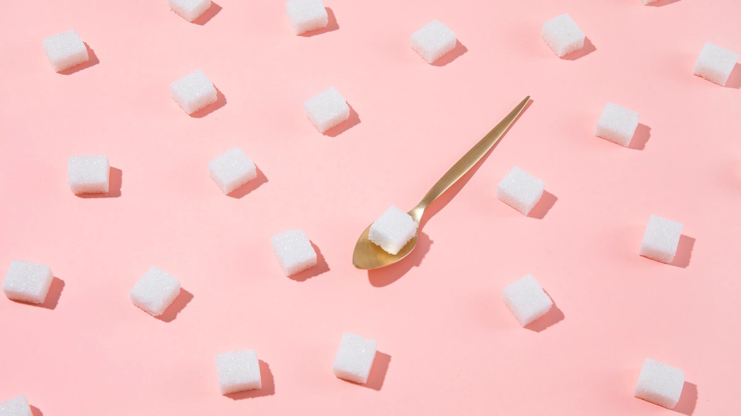 Sugar cubes making a diagonal pattern on a pink background. There is one in the center on a gold spoon.