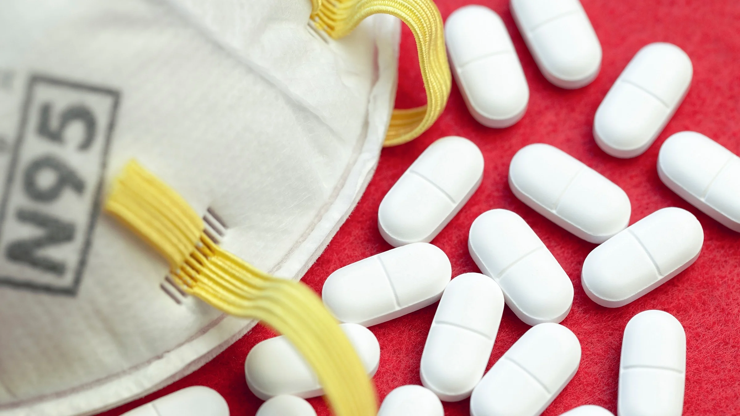 Close-up on a pile of white oblong pills and a N95 mask on a red background.
