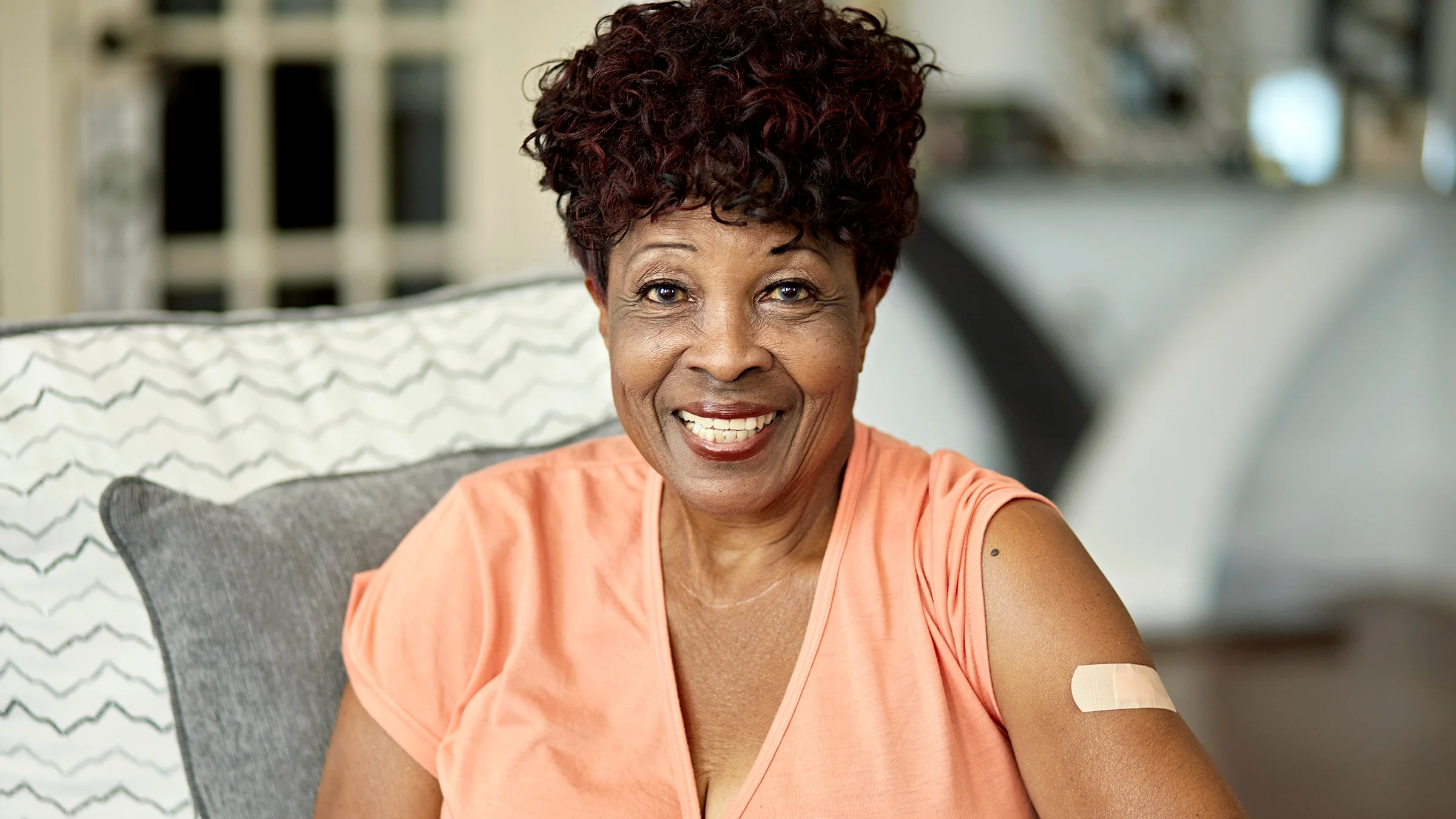 A smiling senior woman with a vaccination bandage on her arm sits on a sofa.