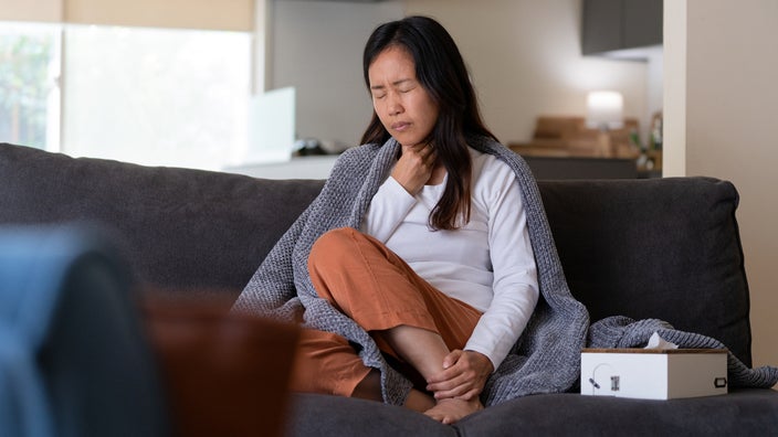 A woman, sitting on the sofa, suffers from throat pain.