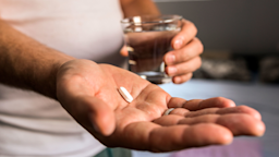 Close-up of a man’s hand. He is holding a white pill and water glass at home.
ljubaphoto/iStock via Getty Images Plus
