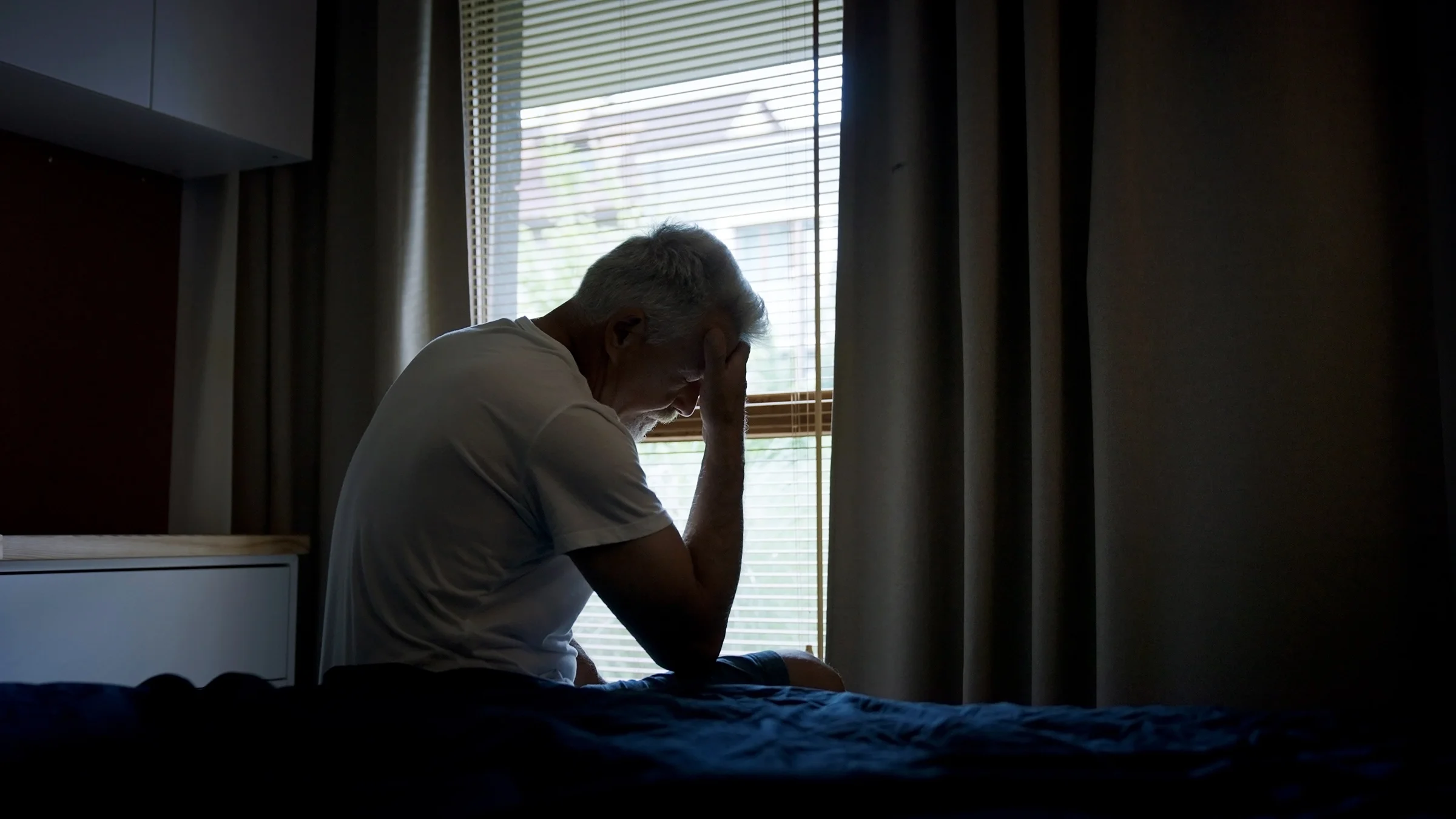 A worried man sits in a dark room with his head in his hands.