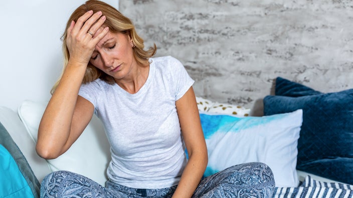 A woman sits in bed and touches her forehead as she experiences a headache. 