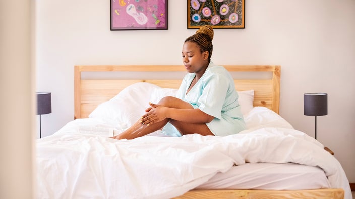 Young woman sitting up in bed and applying lotion to her legs.