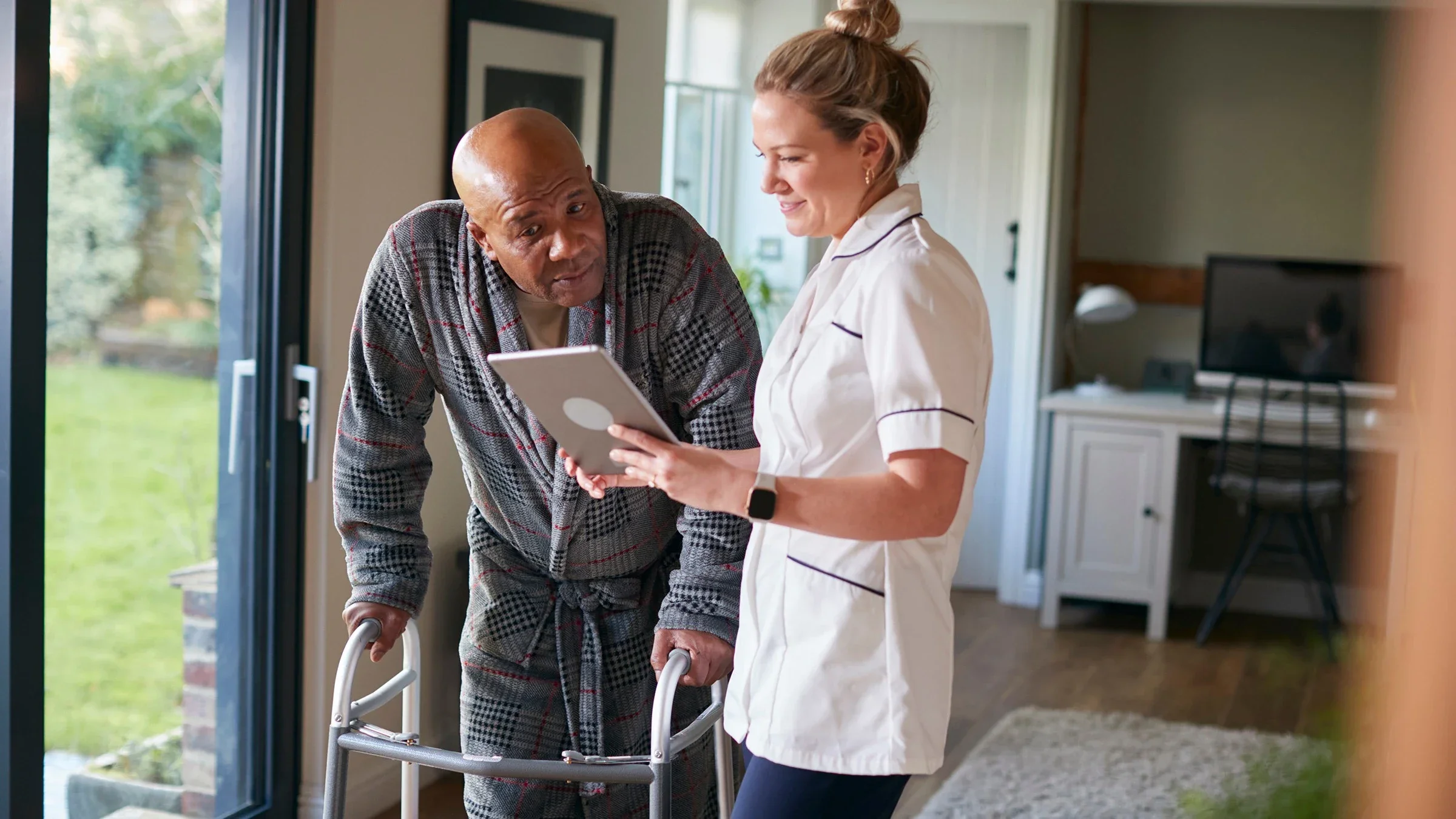 Senior man in robe with his walker talking with a nurse at his home visit. They are reviewing something on a tablet device while standing up.
monkeybusinessimages/iStock via Getty Images