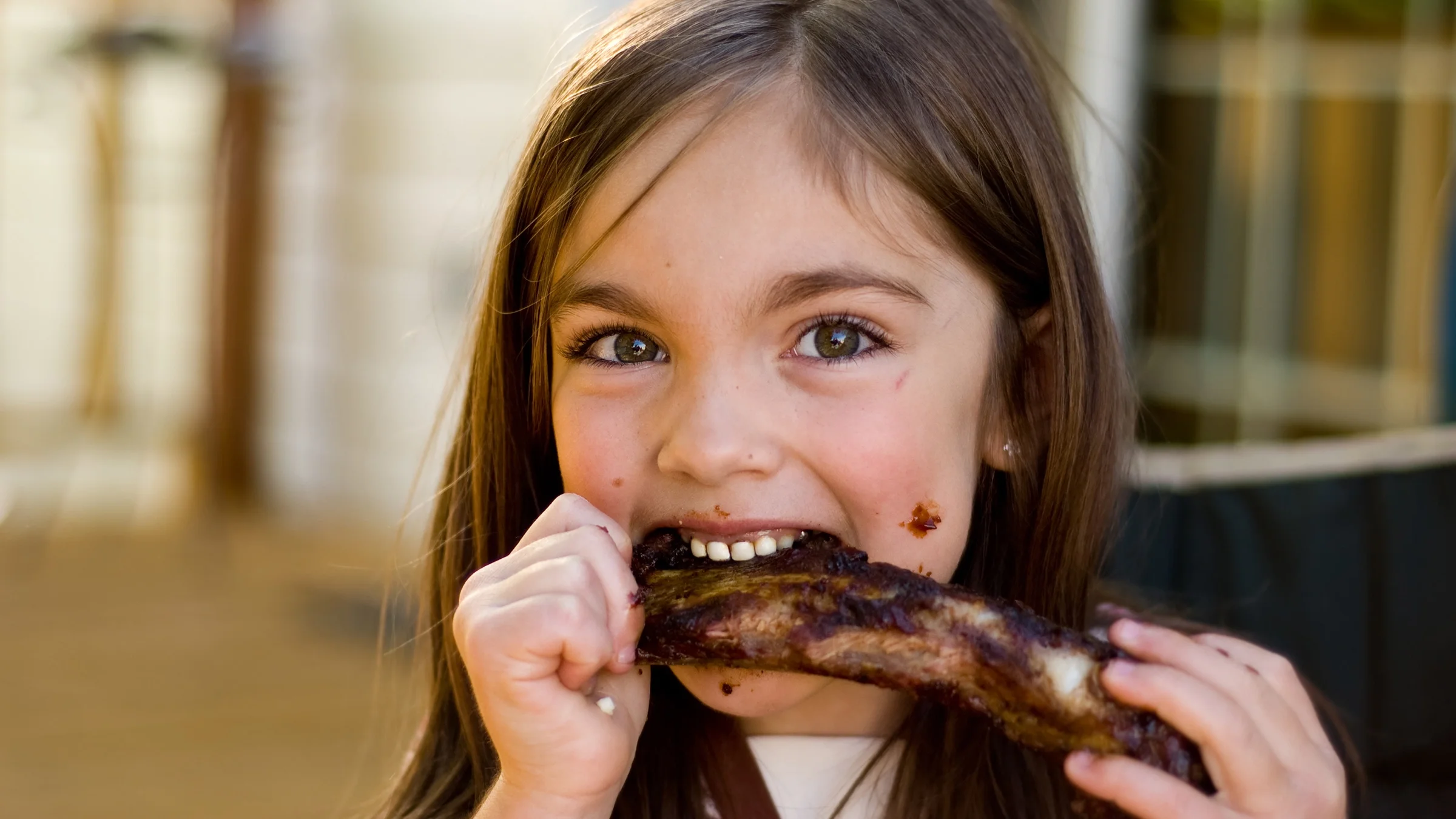 Close-up of a little girl with big brown eyes gnawing on a BBQ rib.